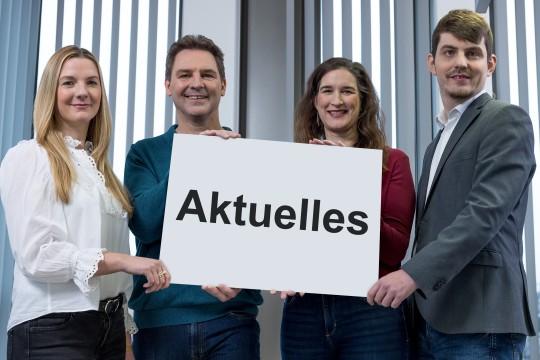 Vier InfoKom-Mitarbeitende halten ein Schild mit der Aufschrift Akuelles