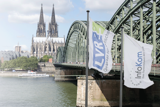 Der Kölner Dom, die Deutzer Brücke und der Rhein mit LVR- und InfoKom-Fahnen ist auf dem Bild zusehen.