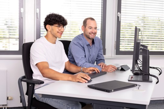 Zwei Männer sitzen an einem Schreibtisch vor einem Computer