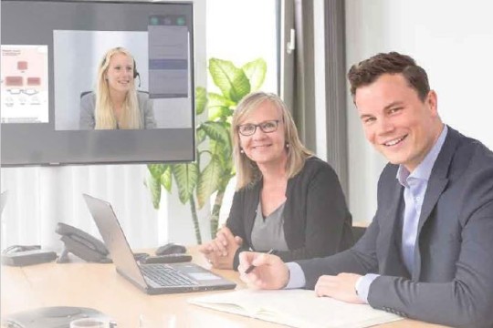 Das Bild zeigt eine Besprechung mit drei Mitarbeitern. Zwei Mitarbeiter (eine Frau und ein Mann) sitzen im Büro und schauen lächelnd in die Kamera. Eine Kollegin nimmt mit Videokonferenz digital teil.