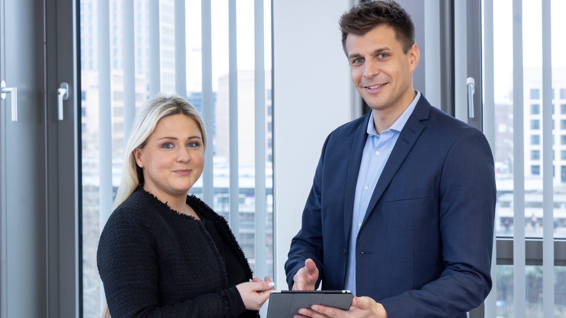 Eine Frau und ein Mann stehen vor einem Fenster und blicken in die Kamera. Der Mann hält ein Tablet in der Hand.