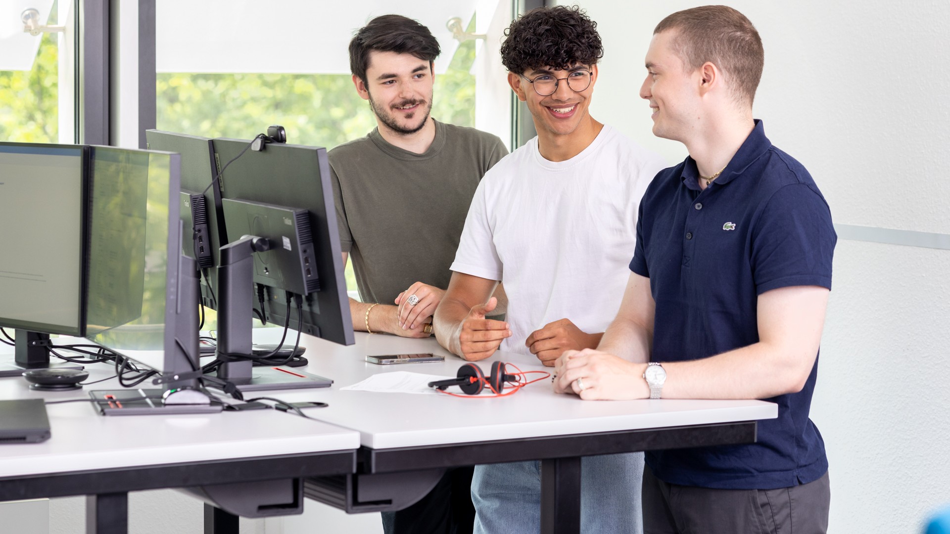 Drei junge Männer stehen an einem höhenverstellbaren Schreibtisch.